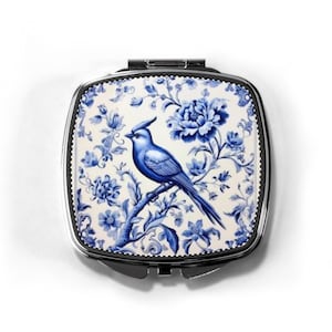 Può includere: Un specchietto compatto quadrato argentato con un design floreale blu e bianco. Il design presenta un uccello blu appollaiato su un ramo, circondato da fiori e foglie blu. Lo specchio ha un'apertura a cerniera.