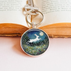 May include: A round silver keychain with a whimsical illustration of a white rabbit leaping across a field under a full moon. The background features a starry night sky and colorful wildflowers. The keychain is attached to a silver key.