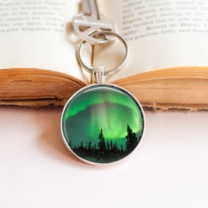 May include: A silver key chain with a round glass charm featuring a green and blue aurora borealis over a dark forest silhouette.
