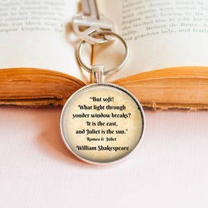 May include: A round silver keychain pendant with a quote from Shakespeare's Romeo & Juliet. The quote is in black text on a cream background. A silver key is attached to the keychain, resting on an open book.