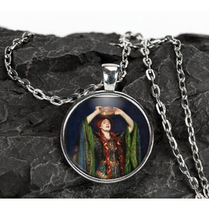 May include: A silver necklace with a round pendant featuring a painting of a woman in a green and gold dress holding a crown above her head. The background is a dark blue.