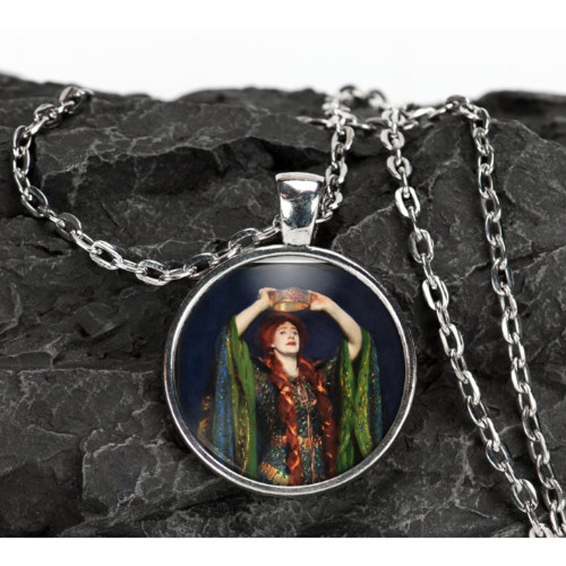 May include: A silver necklace with a round pendant featuring a painting of a woman in a green and gold dress holding a crown above her head. The background is a dark blue.