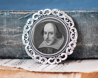 May include: A silver-toned brooch with an ornate design features a black and white portrait of William Shakespeare. The portrait is set in a round glass cabochon.