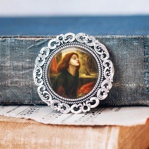 May include: A silver-toned brooch with an ornate frame and a glass cameo featuring a painting of a woman with long red hair looking down. The painting is set against a background of a window with a bird flying outside.