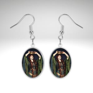 Peut inclure: Une paire de boucles d'oreilles pendantes argentées. Chaque boucle d'oreille présente un pendentif ovale avec une peinture d'une femme en robe verte et bleue, tenant une couronne dorée. La femme a de longs cheveux roux. Les boucles sont suspendues à un crochet.
