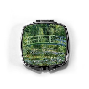 May include: A square, silver-toned compact mirror featuring a painting of a green bridge over a pond filled with lily pads. The artwork is reminiscent of Impressionist art, with a focus on natural light and water reflections.