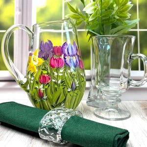May include: A clear glass pitcher hand-painted with purple, pink, and yellow flowers and green foliage, a clear glass mug with a fluted design, and a dark green fabric napkin secured with a clear, faceted, diamond-patterned glass napkin ring.