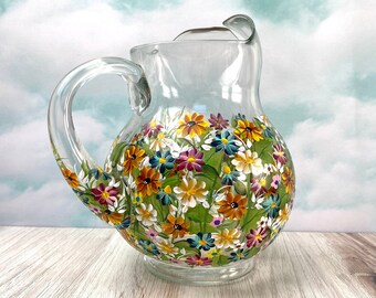 May include: Clear glass pitcher with a rounded body and a handle. The pitcher is decorated with hand-painted colorful flowers in shades of yellow, orange, pink, and blue against a green background. The pitcher sits on a wooden surface.
