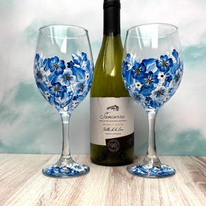 May include: Two clear glass wine glasses featuring hand-painted blue and white floral designs on the bowls and bases, positioned on either side of a Sancerre Pinot Noir wine bottle with a black cap and a label displaying the text Sancerre.