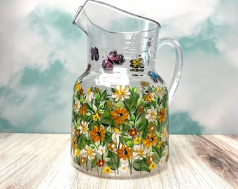 May include: A clear glass pitcher with a floral design. The pitcher is decorated with hand-painted flowers in shades of yellow, orange, white, and green. There are also butterflies painted on the pitcher in shades of purple, blue, and yellow.