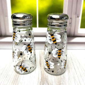 May include: Two clear glass salt and pepper shakers with silver lids. The shakers are decorated with a white and yellow bee design.