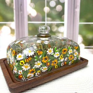 May include: A rectangular wooden serving tray with a clear glass dome. The dome is decorated with hand-painted yellow, white, and orange flowers and green leaves. A clear glass knob sits atop the dome.