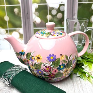 May include: A pink ceramic teapot with a floral design. The teapot has a rounded body, a curved spout, and a handle. The lid is also decorated with flowers. A green napkin and a glass cup are also in the image.