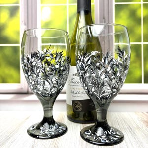 May include: Two wine glasses with a floral design in black and white, and a wine bottle. The glasses have a gray stem and base. The wine bottle has a white label with black text.