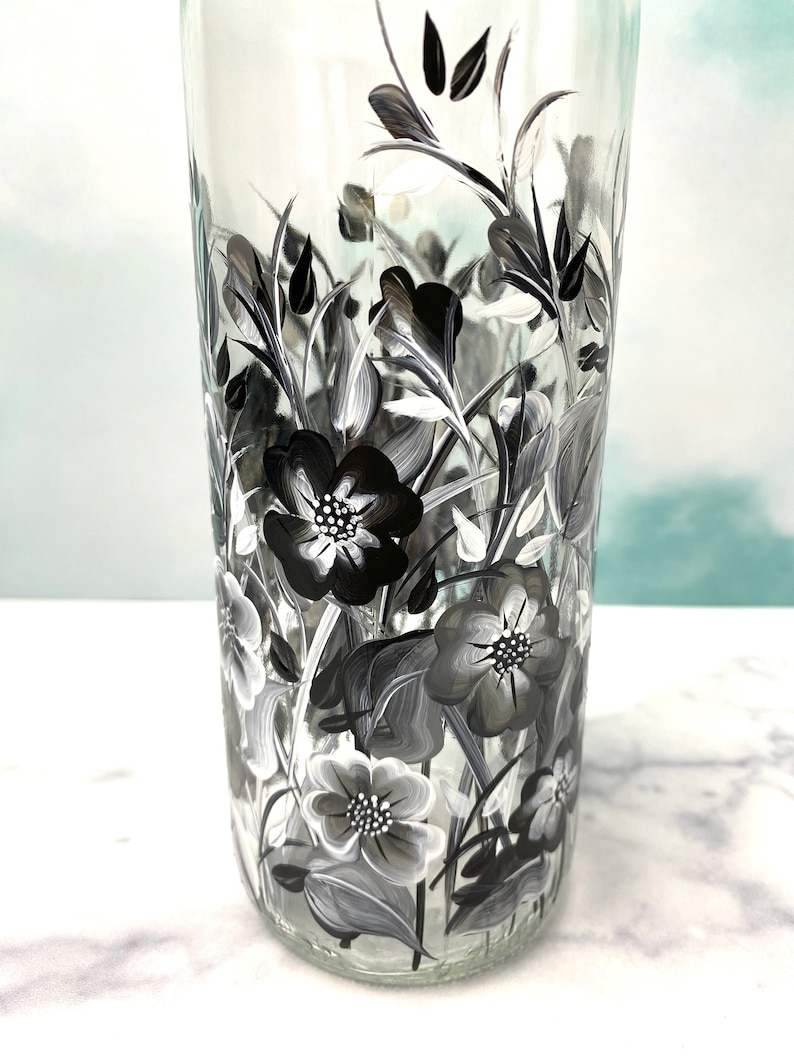 K&ouml;nnte beinhalten: Eine klare Glasflasche mit einem schwarz-wei&szlig;en Blumenmuster, das auf die Seite gemalt ist. Die Blumen sind stilisiert und haben einen Vintage-Look.