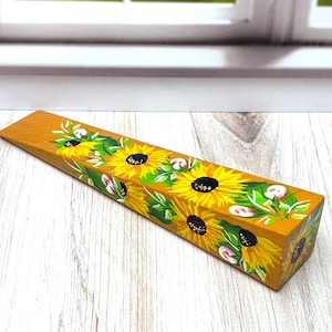 May include: A wedge-shaped wooden doorstop featuring a hand-painted design of bright yellow sunflowers with dark brown centers and green leaves against a warm golden-yellow background, extending along the top surface of the tapered object.