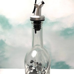 K&ouml;nnte beinhalten: Ein silberner und schwarzer Flaschenausgie&szlig;er mit einem Hebel oben. Der Ausgie&szlig;er ist an einer durchsichtigen Glasflasche mit einem schwarz-wei&szlig;en Blumendesign befestigt.