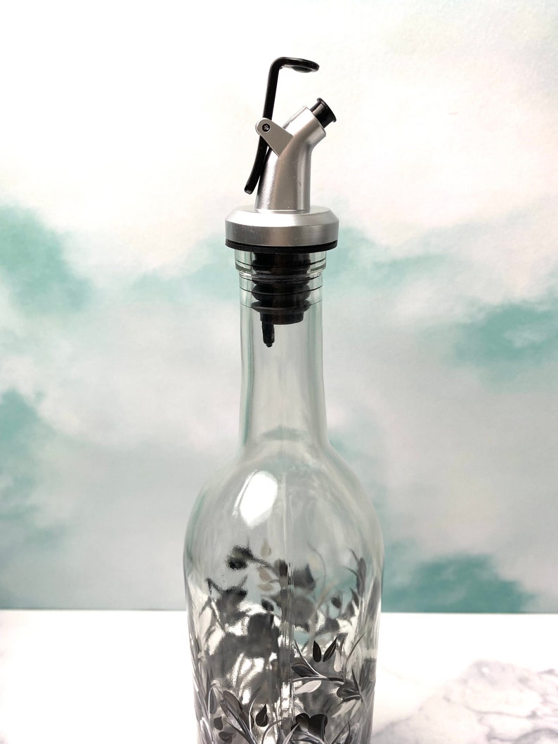 K&ouml;nnte beinhalten: Ein silberner und schwarzer Flaschenausgie&szlig;er mit einem Hebel oben. Der Ausgie&szlig;er ist an einer durchsichtigen Glasflasche mit einem schwarz-wei&szlig;en Blumendesign befestigt.