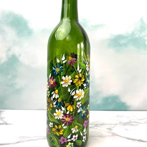 May include: A green glass bottle hand-painted with a colorful floral design. The bottle is decorated with a variety of flowers, including daisies, wildflowers, and other blooms in shades of pink, yellow, white, blue, and green.