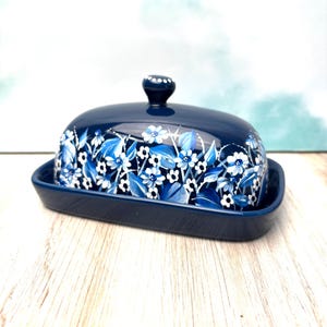 May include: A navy blue ceramic butter dish with a matching lid. The lid and base are adorned with a floral pattern in shades of blue and white. The lid has a small knob on top. The dish is rectangular.