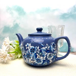 May include: A blue ceramic teapot with a floral design in shades of blue and white. The teapot has a lid, spout, and handle. The background includes a glass and white flowers.