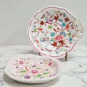 May include: Two white porcelain serving dishes with floral patterns. The larger dish has a scalloped edge and a handle. The smaller dish has a more rounded shape. Both dishes feature intricate floral designs in shades of pink, blue, green, and brown.