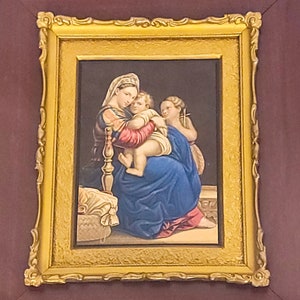 May include: A framed painting depicting the Madonna and Child with a cherub. The painting is in a gold frame and features a mother holding a child in her arms. The cherub is holding a cross. The painting is set against a dark background.