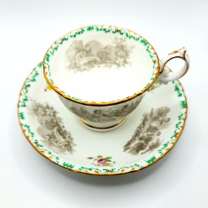 May include: A white teacup and saucer with a green and gold trim. The cup and saucer feature a black and white pastoral scene with a floral motif.