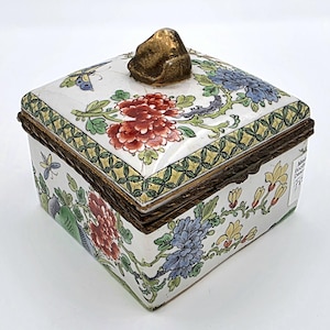 May include: A square, white porcelain trinket box with a hinged lid. The box is decorated with colourful floral and butterfly designs. The lid features a gold-coloured knob. The box has a dark brown border.