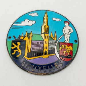 Puede incluir: Un pin redondo de metal con un diseño colorido que representa un edificio con una aguja, una figura orinando y el texto "BRUXELLES".