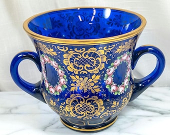 Op de afbeelding: Een kobaltblauwe decoratieve vaas met twee handvatten, met gouden filigraan en bloemmotieven. De vaas heeft een gouden rand en is versierd met roze en rode rozen. Het ontwerp is sierlijk en elegant.