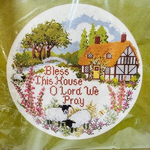 May include: A white cross-stitch embroidery with a cottage, trees, flowers, and sheep. The text reads "Bless This House O Lord We Pray".