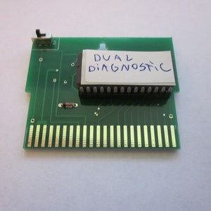 Op de afbeelding: Een groene printplaat met gouden connectoren en een zwarte geïntegreerde schakeling. De tekst "DUAL DIAGNOSTIC" is handgeschreven op een wit label dat aan de printplaat is bevestigd. Een kleine schakelaar bevindt zich in de linkerbovenhoek.