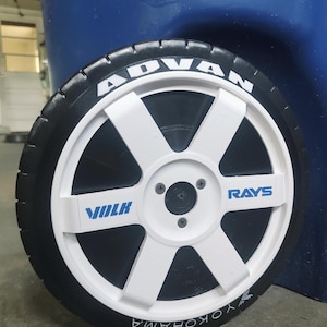 May include: A white six-spoke wheel with a black tire featuring white lettering that reads ADVAN and YOKOHAMA. The wheel center has blue text reading VOLK and RAYS, with a black center cap secured by three visible screws on the white rim face.