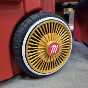 May include: A black rubber wheel with a white sidewall accent attached to a red plastic storage unit. The wheel features a gold-colored, multi-spoke hub design with a central red circular cap displaying a white letter M logo in a stylized font.
