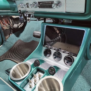 May include: A custom teal automotive center console featuring a rectangular touchscreen display, three circular gauges, two stainless steel cup holders, and a row of metal toggle switches. The console has a silver engine-turned metal trim.