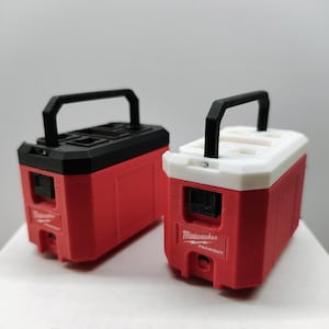 May include: Two red and black miniature toolboxes with black handles. One has a black top, the other a white top. The text "Milwaukee PACKOUT" is visible on the front of the red toolboxes. The toolboxes are on a white surface.