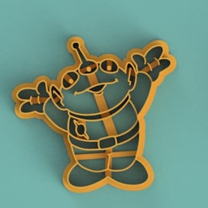 May include: A yellow cookie cutter shaped like a cartoon alien with three eyes, a big smile, and outstretched arms. The alien is wearing a belt and has a small circle on its chest.