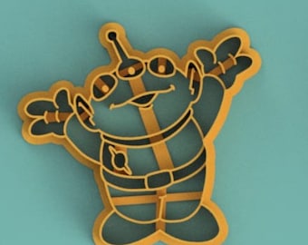 May include: A yellow cookie cutter shaped like a cartoon alien with three eyes, a big smile, and outstretched arms. The alien is wearing a belt and has a small circle on its chest.