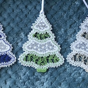 May include: Five Christmas tree ornaments with white lace detailing and colourful accents. Each tree has a different colour scheme: gold, navy blue, green, grey, and gold. They are hanging from white ribbons, perfect for holiday decor.