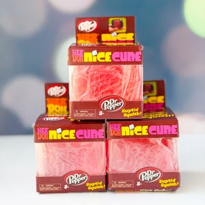 Op de afbeelding: Gestapelde dozen met "Nee Doh Nice Cube" speelgoed, met een Dr. Pepper logo. Elke bruine doos met roze en gele tekst bevat een rode, squishy kubus. De dozen zijn gestapeld op een wit oppervlak tegen een vage achtergrond.