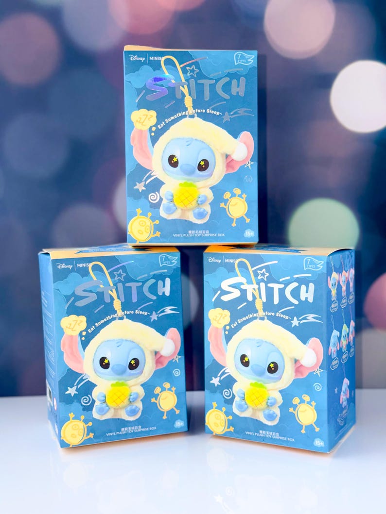 May include: Three blue boxes featuring a Stitch character in a yellow outfit holding a pineapple. The boxes have the word "STITCH" in a stylized font and the phrase "Eat Something before Sleep". The boxes are on a white surface.