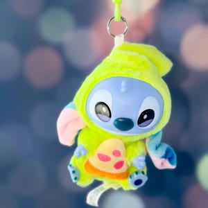 May include: A plush Stitch toy in a lime green hooded outfit with a pizza slice design. The toy has blue skin, large black eyes, and pink and blue accents. It hangs from a green lanyard.