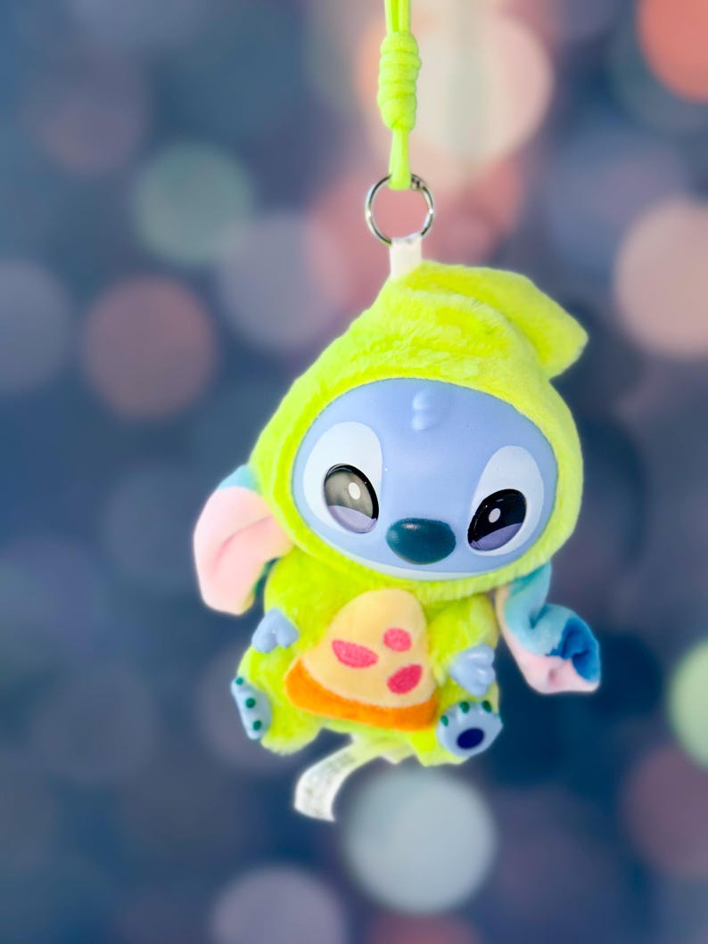 May include: A plush Stitch toy in a lime green hooded outfit with a pizza slice design. The toy has blue skin, large black eyes, and pink and blue accents. It hangs from a green lanyard.