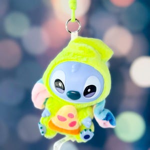 May include: A plush Stitch character in a lime green hooded outfit with a pizza slice design. The blue-skinned alien has large black eyes and a green nose. A lime green lanyard is attached to the plush toy.