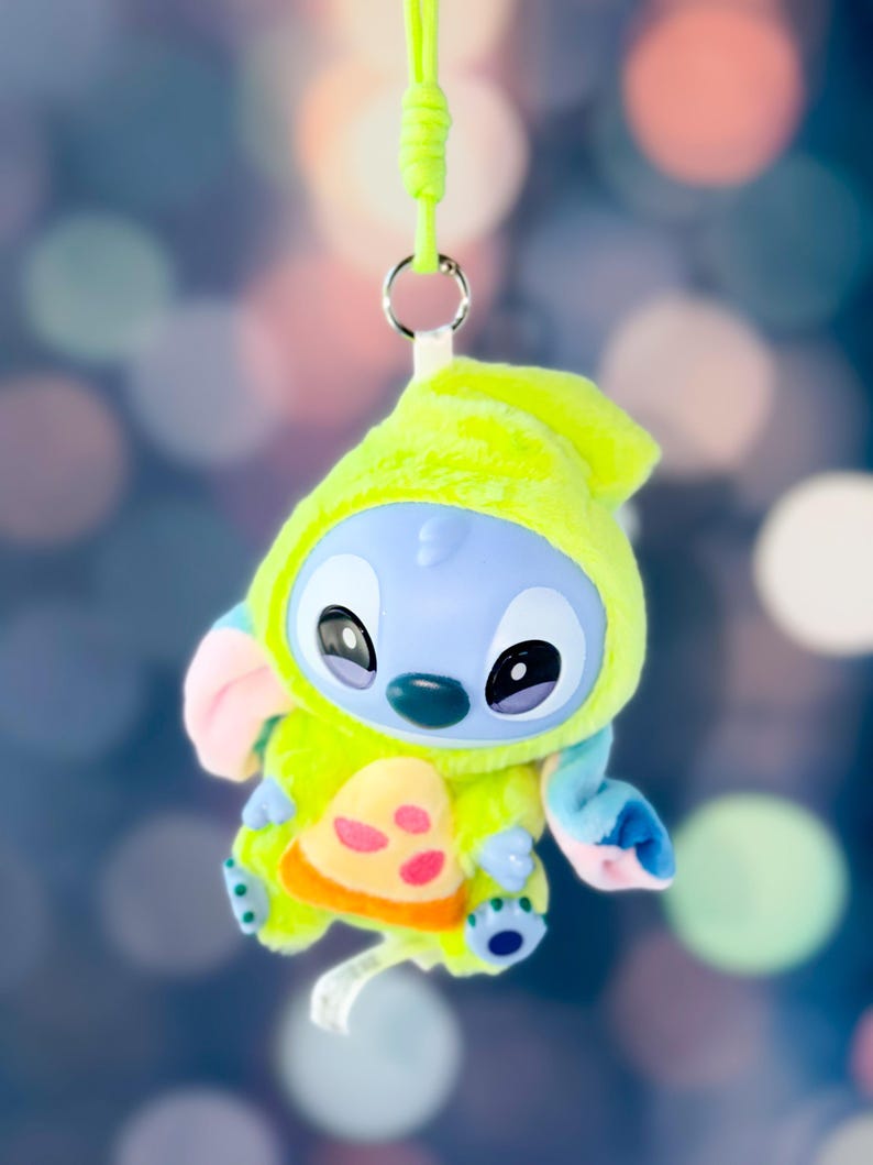 May include: A plush Stitch character in a lime green hooded outfit with a pizza slice design. The blue-skinned alien has large black eyes and a green nose. A lime green lanyard is attached to the plush toy.