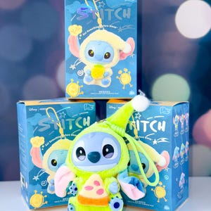 May include: A blue plush Stitch toy wearing a lime green outfit and holding a pizza slice, with a matching box in the background. The box has the word "Stitch" and the phrase "Eat Something Before Sleep".