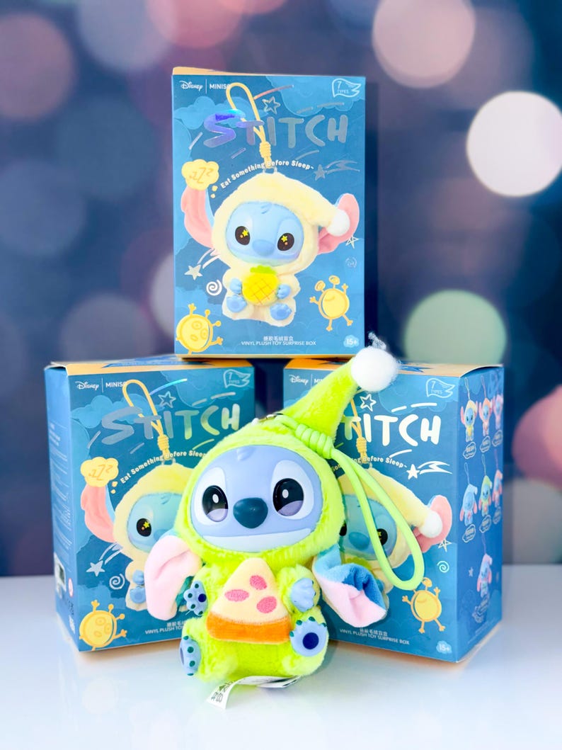 May include: A blue plush Stitch toy wearing a lime green outfit and holding a pizza slice, with a matching box in the background. The box has the word "Stitch" and the phrase "Eat Something Before Sleep".