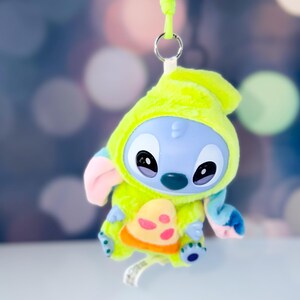 May include: A plush Stitch figure in a lime green hooded outfit with a pizza-shaped design on the front. The figure has blue skin, large black eyes, and pink ears. A lime green lanyard is attached to the top.