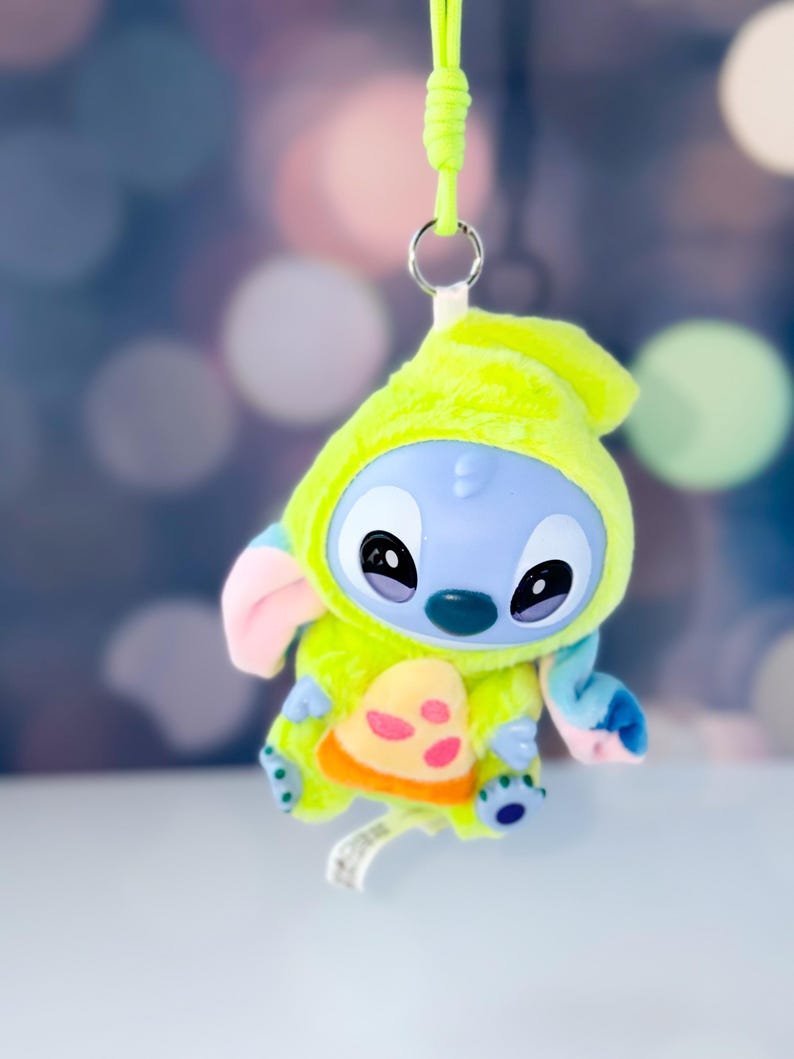 May include: A plush Stitch figure in a lime green hooded outfit with a pizza-shaped design on the front. The figure has blue skin, large black eyes, and pink ears. A lime green lanyard is attached to the top.